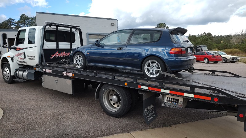 Dark blue hatchback securely loaded on a tow truck in a parking lot, highlighting Anthony’s Towing’s commitment to family road trip safety.