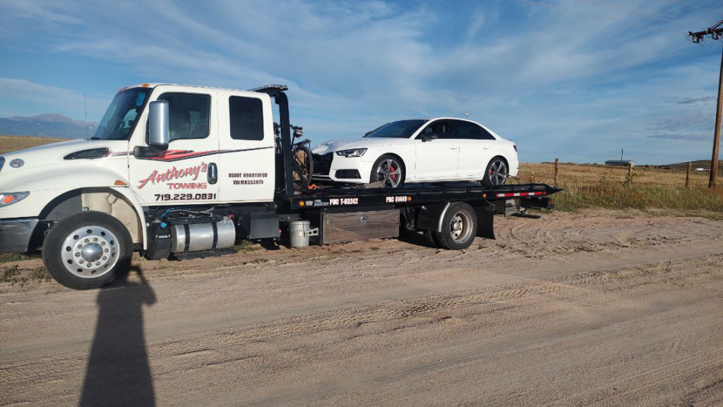 A white sedan, ready for a holiday adventure, gets a lift on a tow truck in an empty lot, with the subtle shadow of an onlooker capturing the moment. Trust Anthony's Towing for reliable service in your holiday road trip planning.