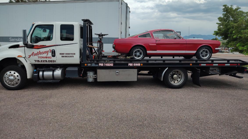 A red car on a tow truck awaits rescue in a lot, with a trailer in the background. Trust Anthony's Towing for dependable service for your holiday road trip planningto go according to schedule.